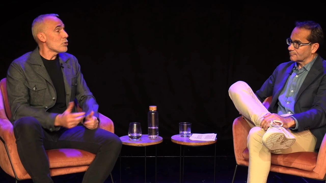 Fotograma del vídeo original: dos personas conversan sentadas en sillones, con dos mesas bajas entre ellas, en un escenario oscuro.